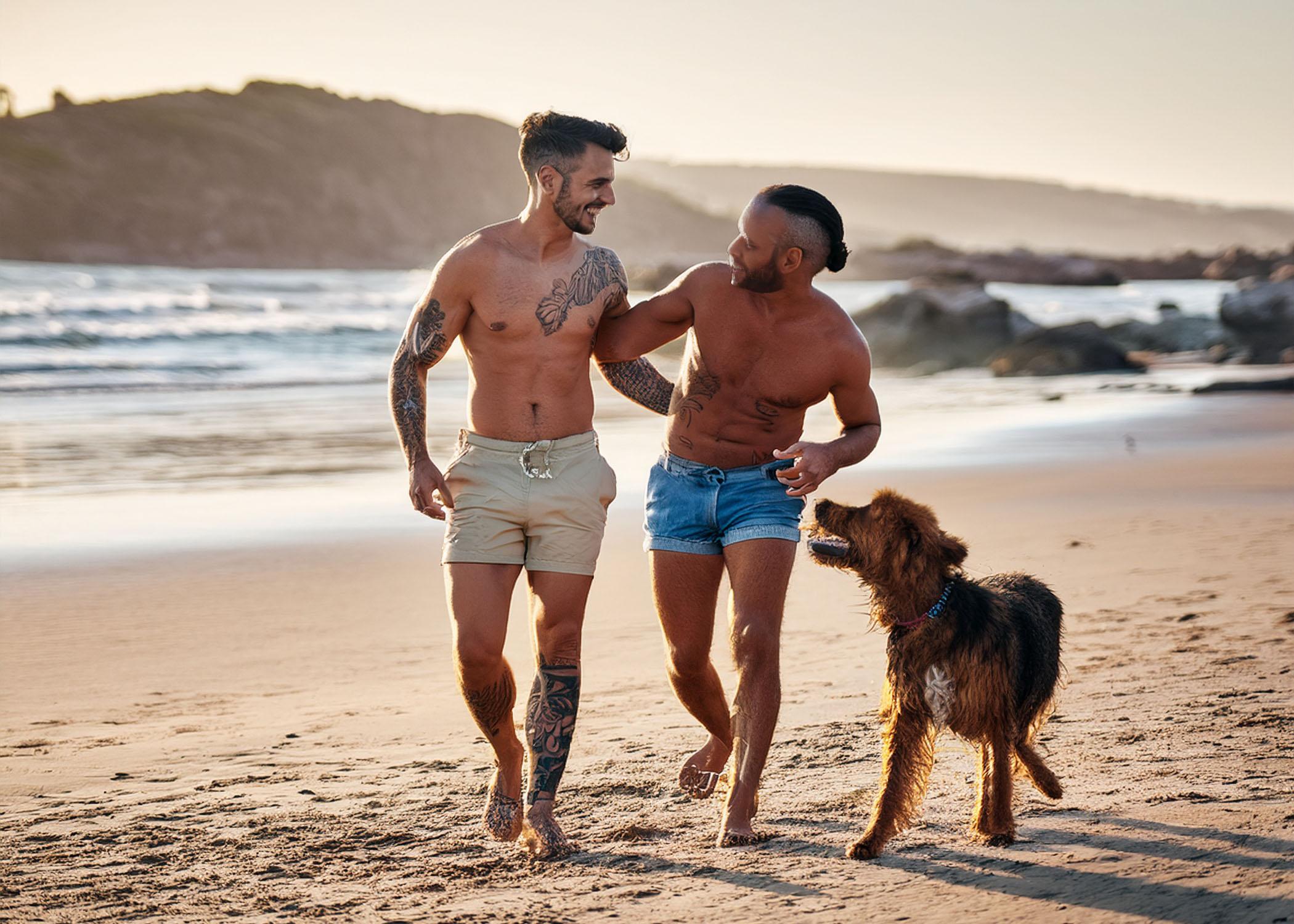 gay couple playing with dog at beach in shorts